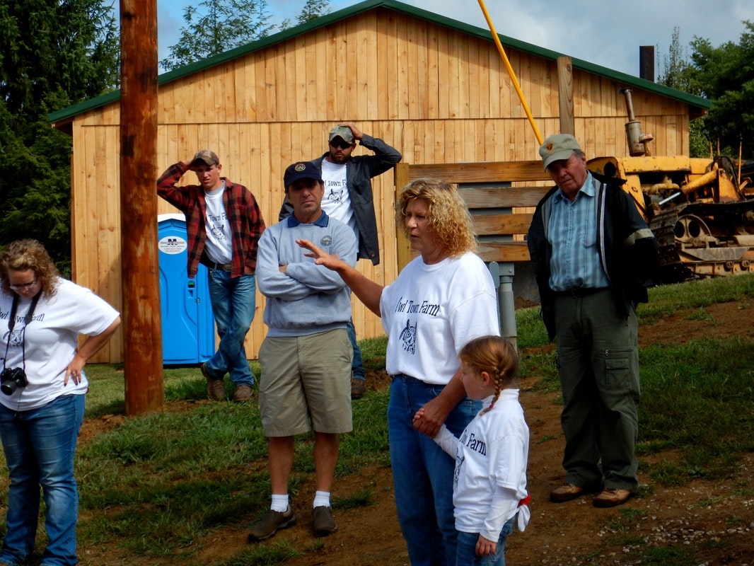 A group of people standing outdoors near a wooden building and a bulldozer. One person gestures while talking, others listen or look around.