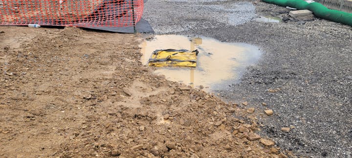 Construction site with gravel, dirt, a puddle, a yellow object, and an orange fence.