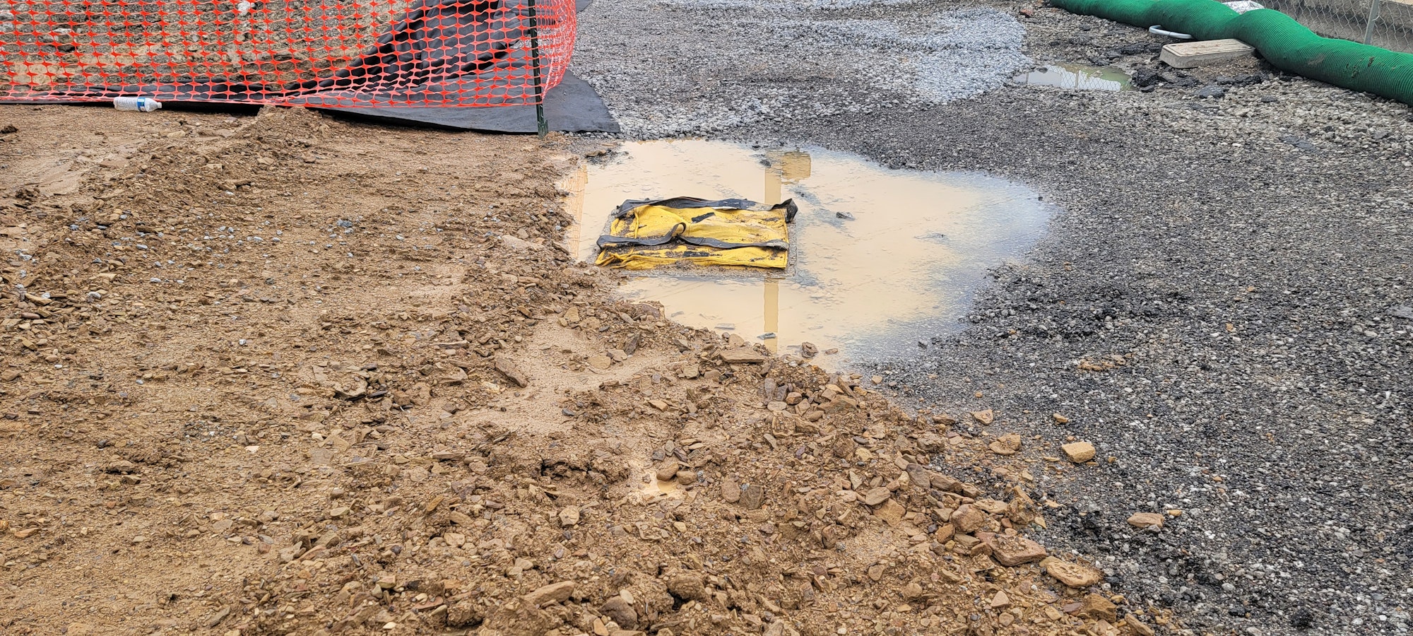 Construction site with gravel, dirt, a puddle, a yellow object, and an orange fence.