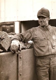 A man in work attire leans on a bin filled with coal, wearing a cap and gloves, appearing dusty and grimy.