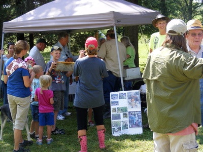 People gathering under a canopy at an outdoor event, with children, posters, and a sunny day.