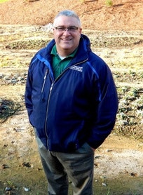 A person wearing a blue jacket and glasses stands outdoors with a smile, hands in pockets, on a sunny day.