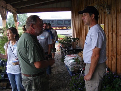 People talking outside near a wooden building, with plants and a bus in the background.