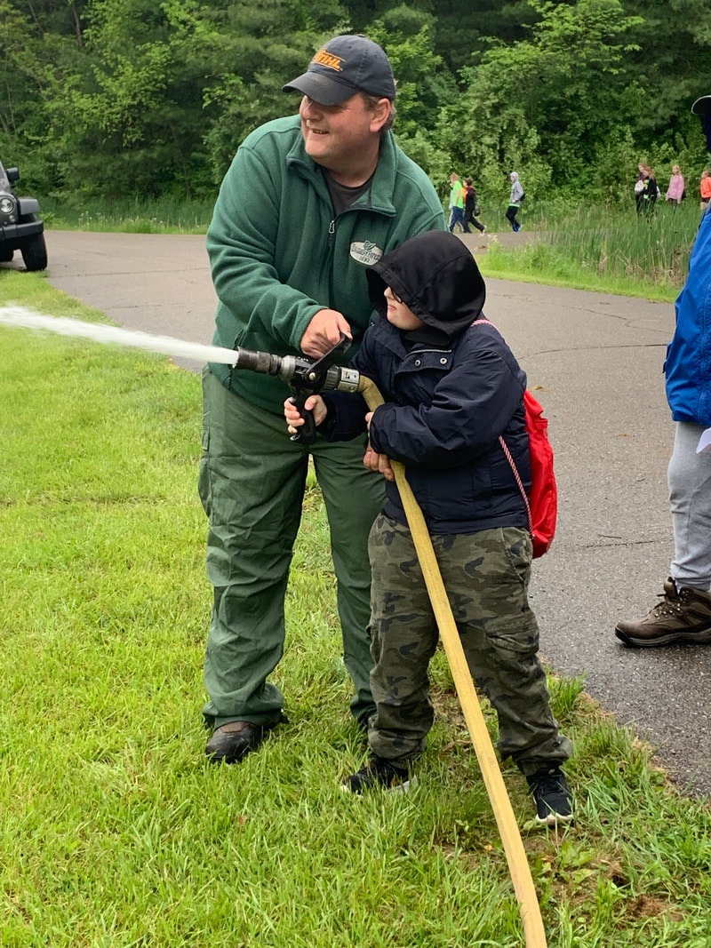 A man is helping a child spray water from a fire hose in a grassy area.