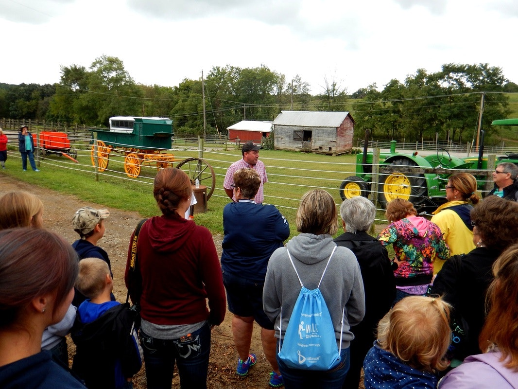 A group of people gathered outdoors, listening to a speaker near vintage vehicles and a tractor in a rural setting.
