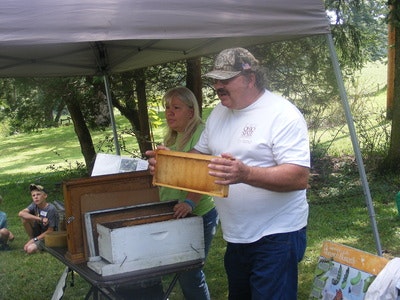 Two people demonstrating beekeeping equipment under a tent outdoors, with an audience in the background.
