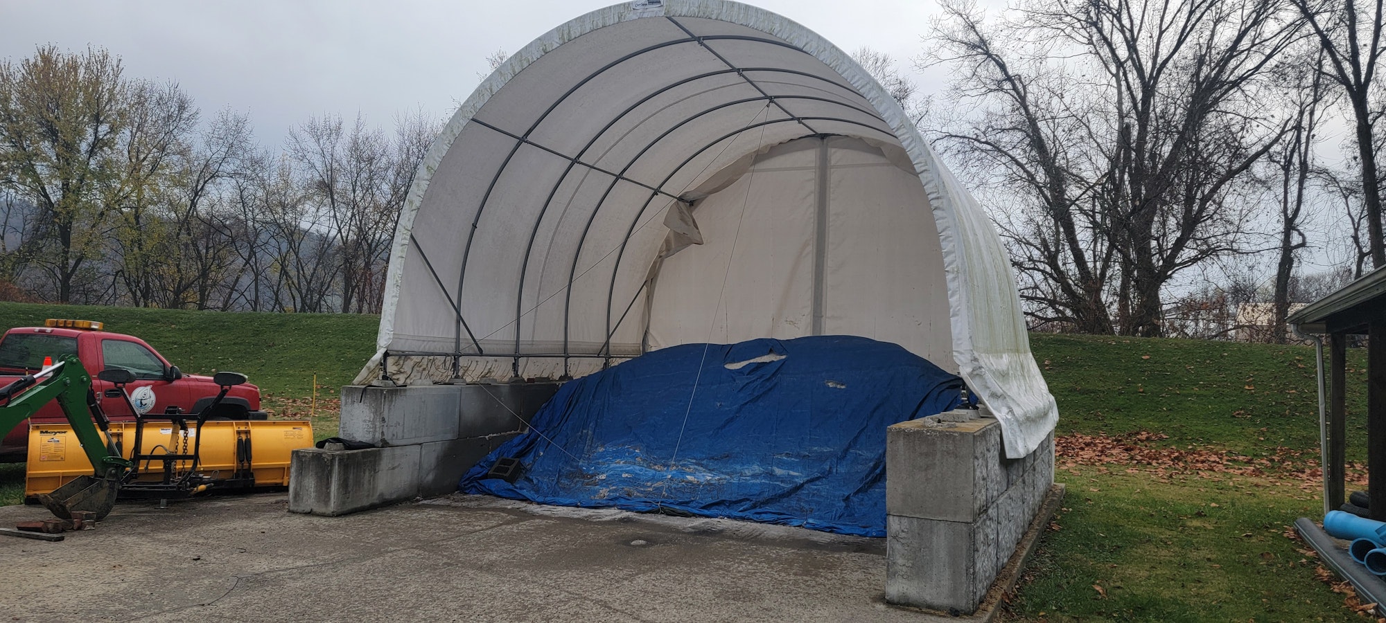 A covered structure storing items under a blue tarp, with a red truck and a yellow snowplow attachment nearby.