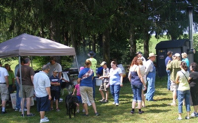 A group of people gathered outdoors, some under a canopy, with trees in the background, possibly at a picnic or event.