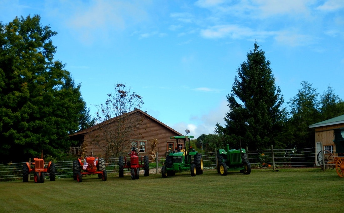 Several tractors lined up on grass with trees and a wooden house in the background.