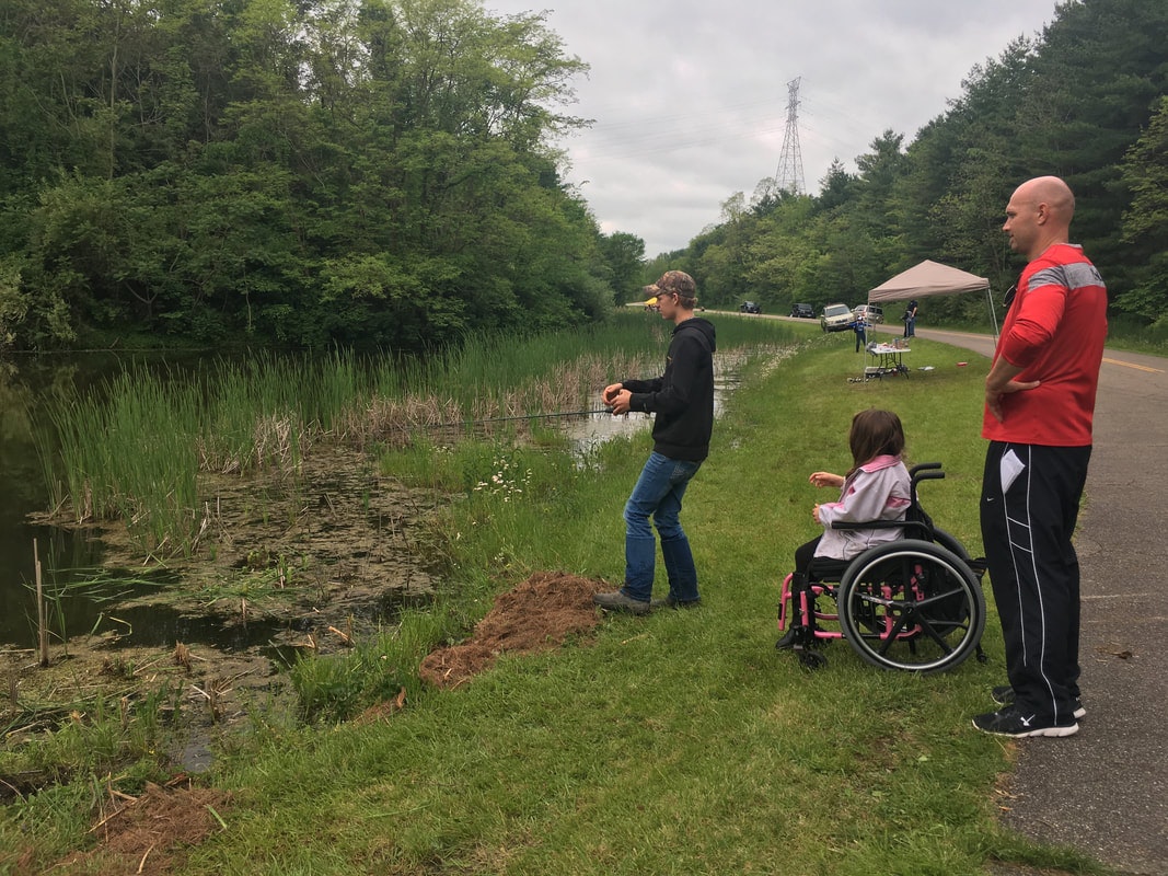 A person fishes by a pond with two others observing, one in a wheelchair, surrounded by greenery and an overcast sky.