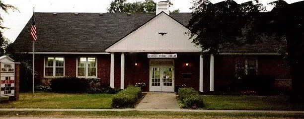 The image shows a brick building that appears to be a public library, featuring a front entrance and well-maintained landscaping.