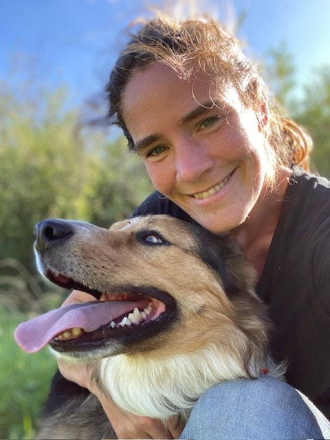 A person smiling while taking a selfie with a happy dog outdoors in a green setting.