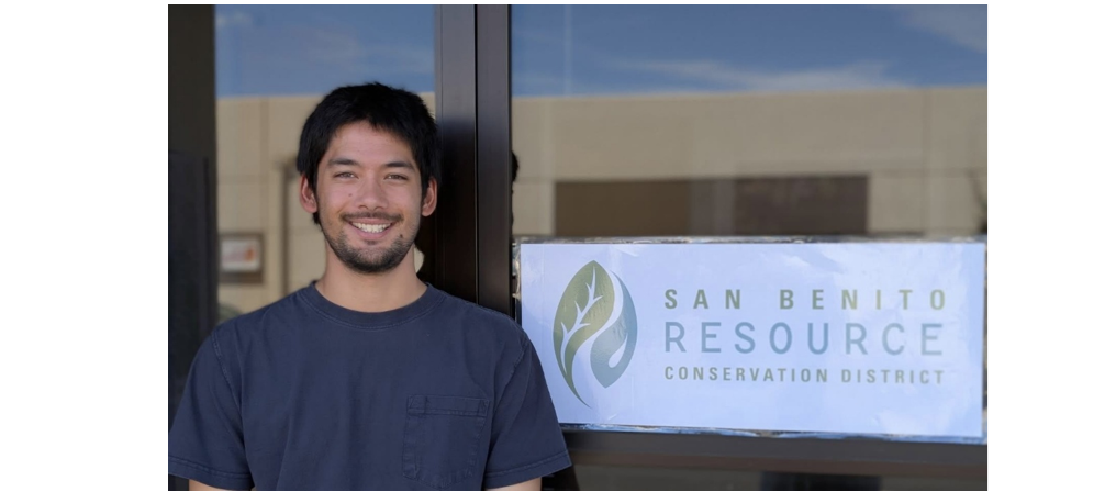 A person smiling next to a sign reading "San Benito Resource Conservation District."