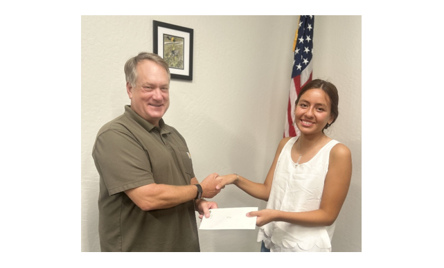 Two people smiling, shaking hands, one holding an envelope, with a flag and framed picture in the background.