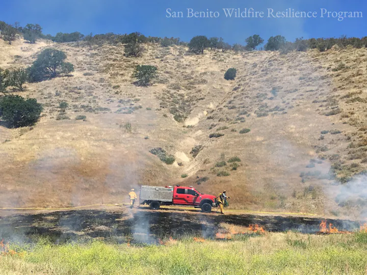 Firefighters and a red truck in a hilly area, managing a small grass fire.