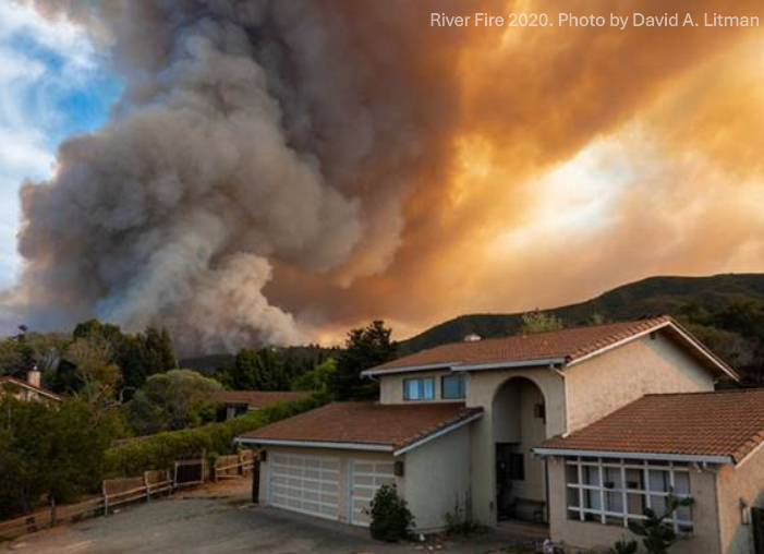 A large plume of smoke rises behind a residential house, indicating a nearby wildfire.