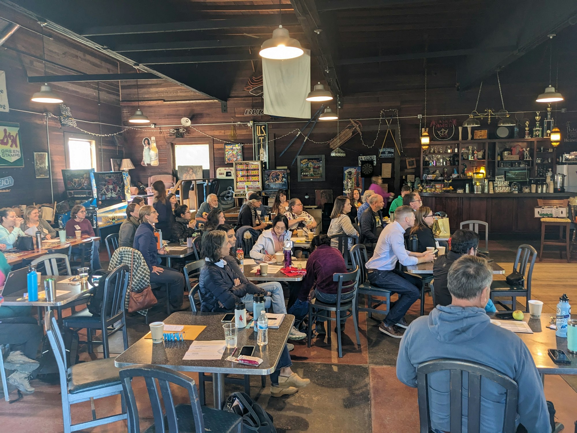A group of people seated in a rustic room, attentively listening, surrounded by tables and a bar area.
