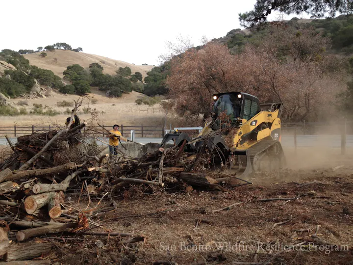 A small bulldozer and workers clearing debris in a rural area with hills and trees in the background.