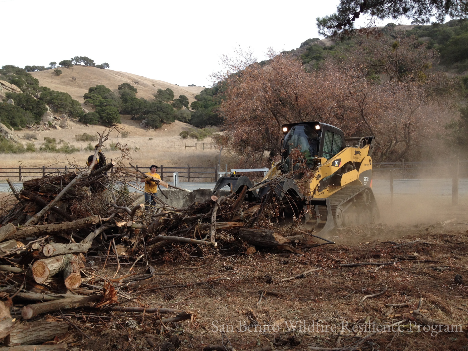 A small bulldozer and workers clearing debris in a rural area with hills and trees in the background.
