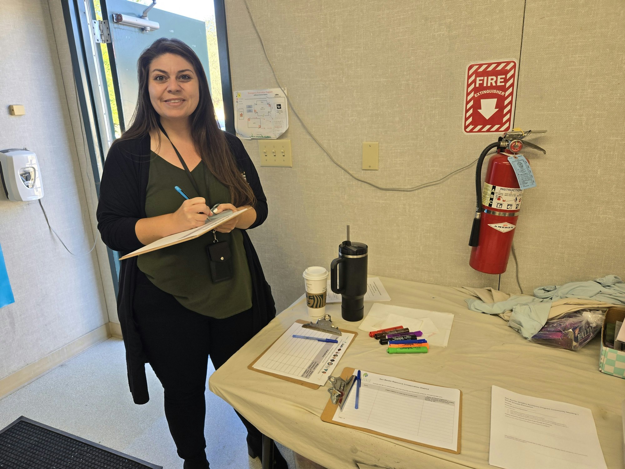 A woman stands by a desk with papers and markers, near a fire extinguisher on the wall. Madison Mitchell, San Benito County Office of Emergency Services
