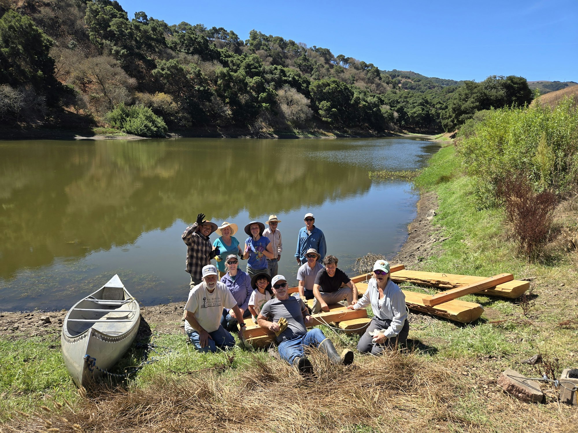 A group of people posing by a lakeside with a canoe and wooden planks, surrounded by greenery and hills.