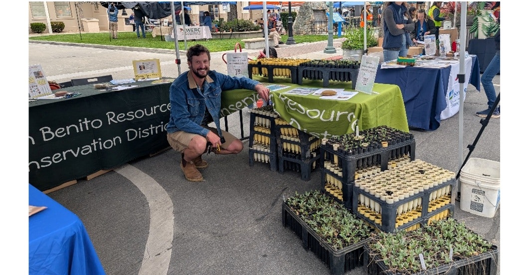 A person squats near plant trays at an outdoor resource conservation booth.