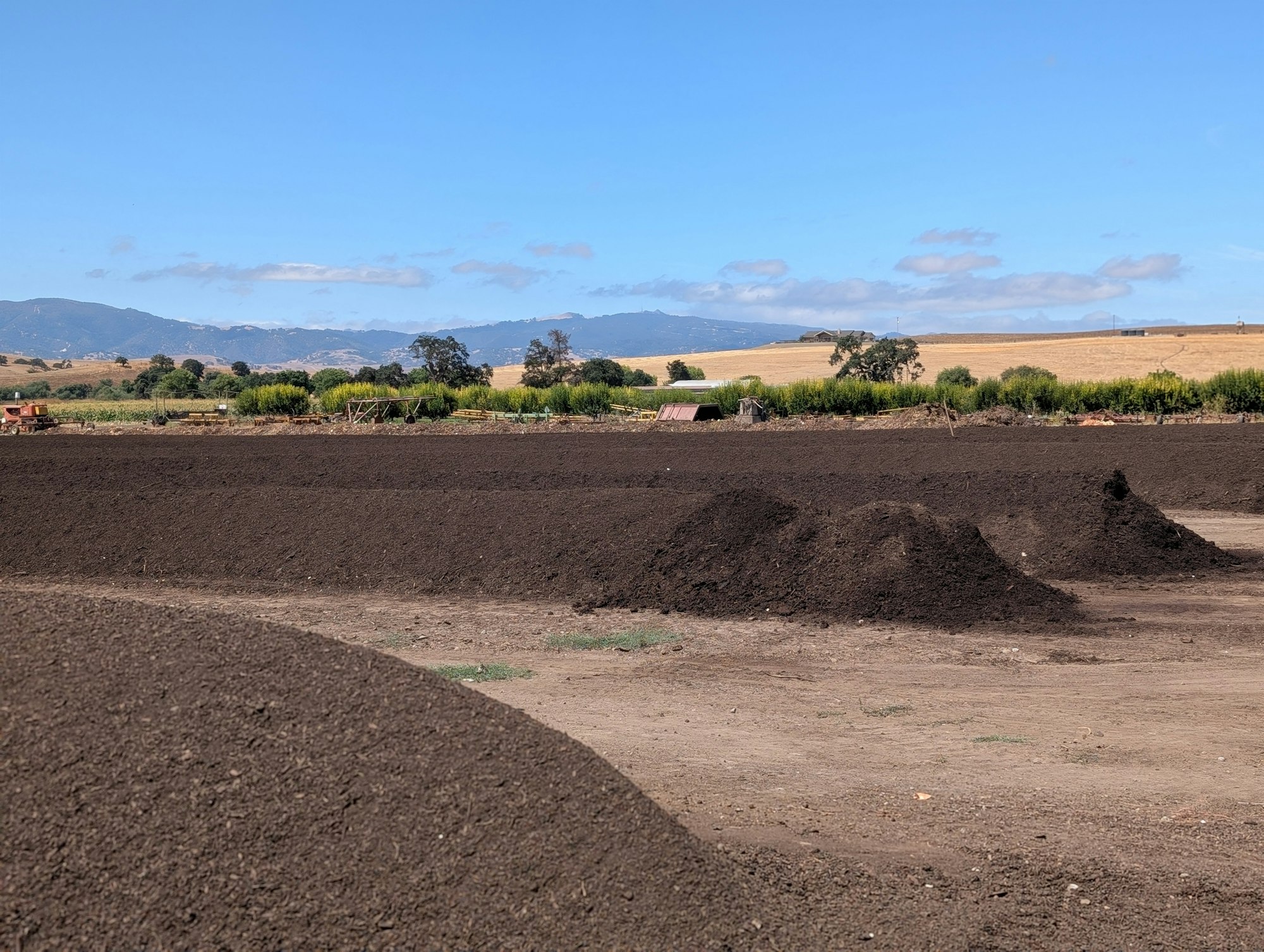 The image shows a landscape with dark mounds of soil or compost under a blue sky and rolling hills in the background.