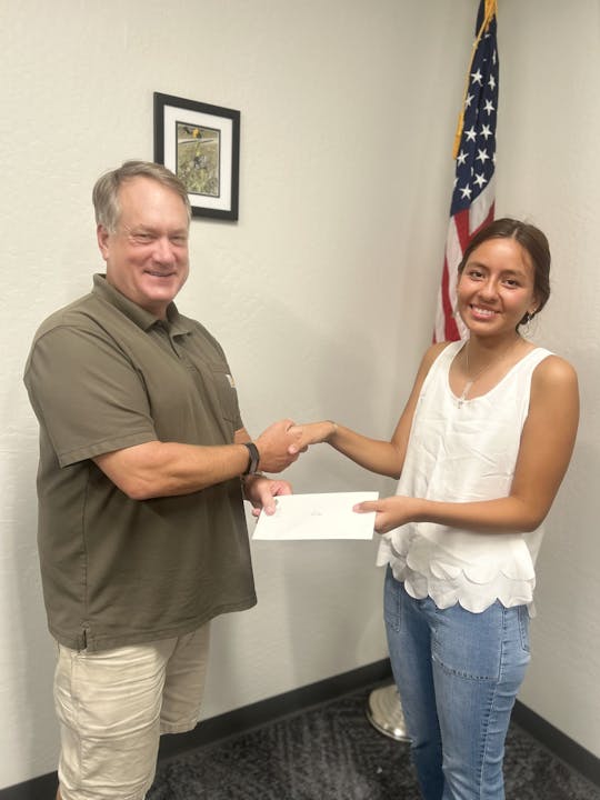 Two people are shaking hands, one handing an envelope to the other, standing near a U.S. flag.