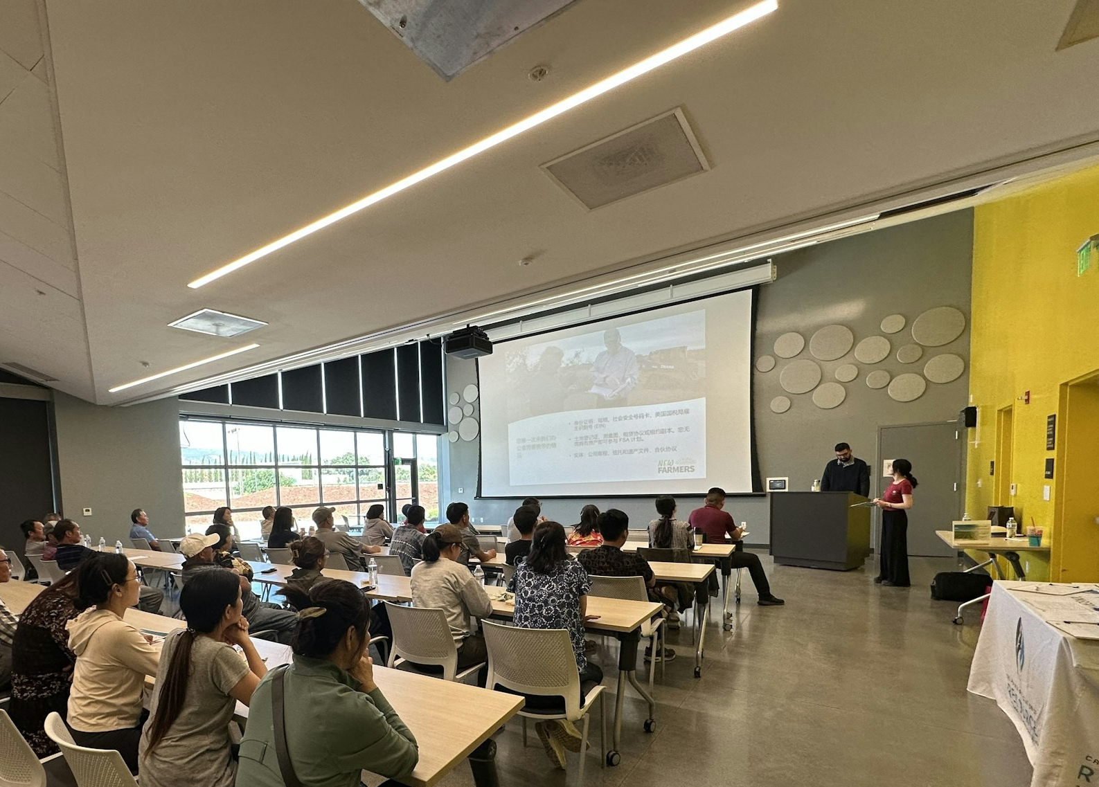 People attending a presentation in a classroom, with a speaker at a podium and a projected screen.