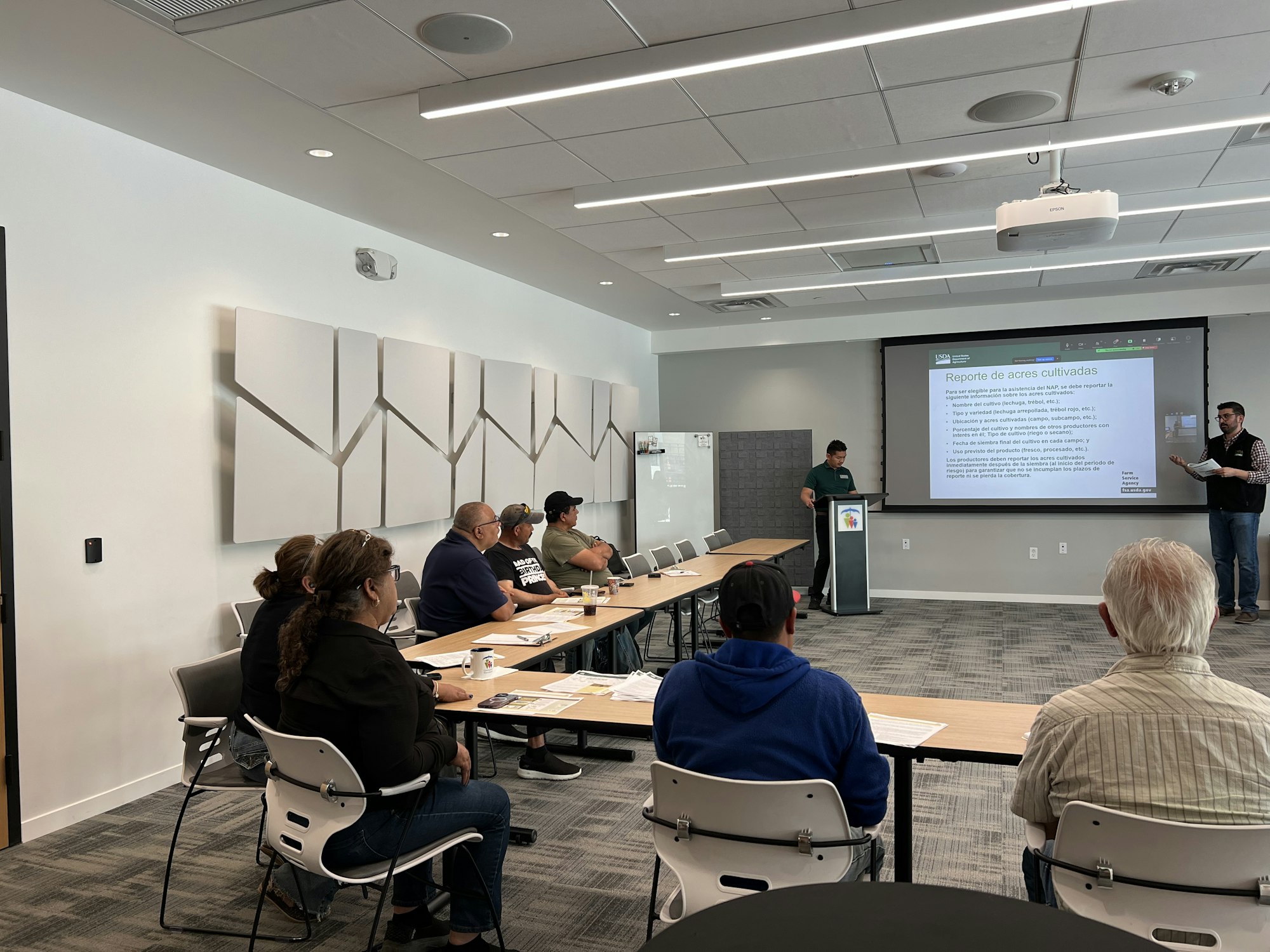 People attend a presentation in a conference room. A speaker presents slides on "Reporte de acres cultivadas" in Spanish.