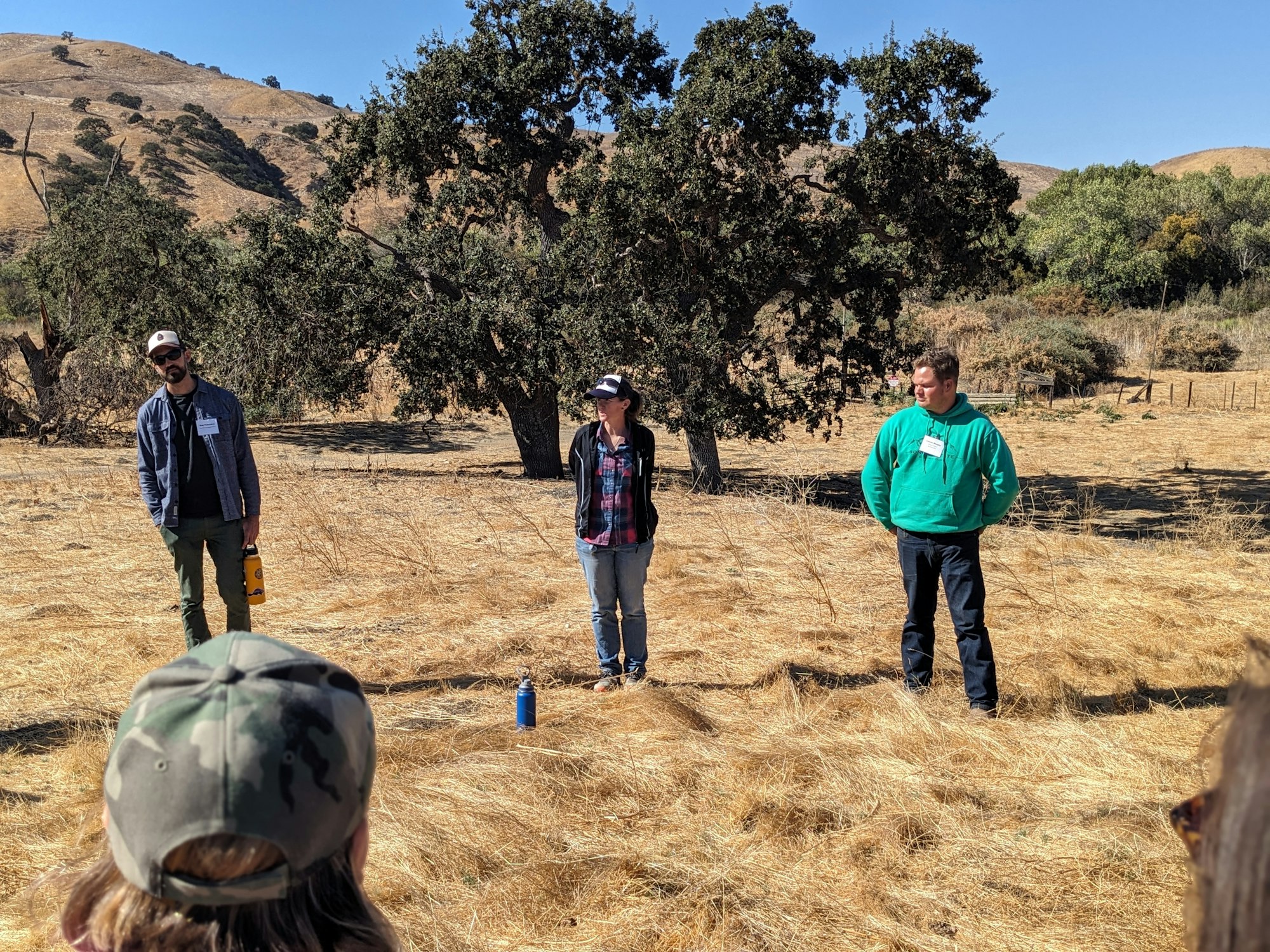 Three people stand in a dry, grassy field with trees and hills in the background. One holds a thermos. Audience in foreground.