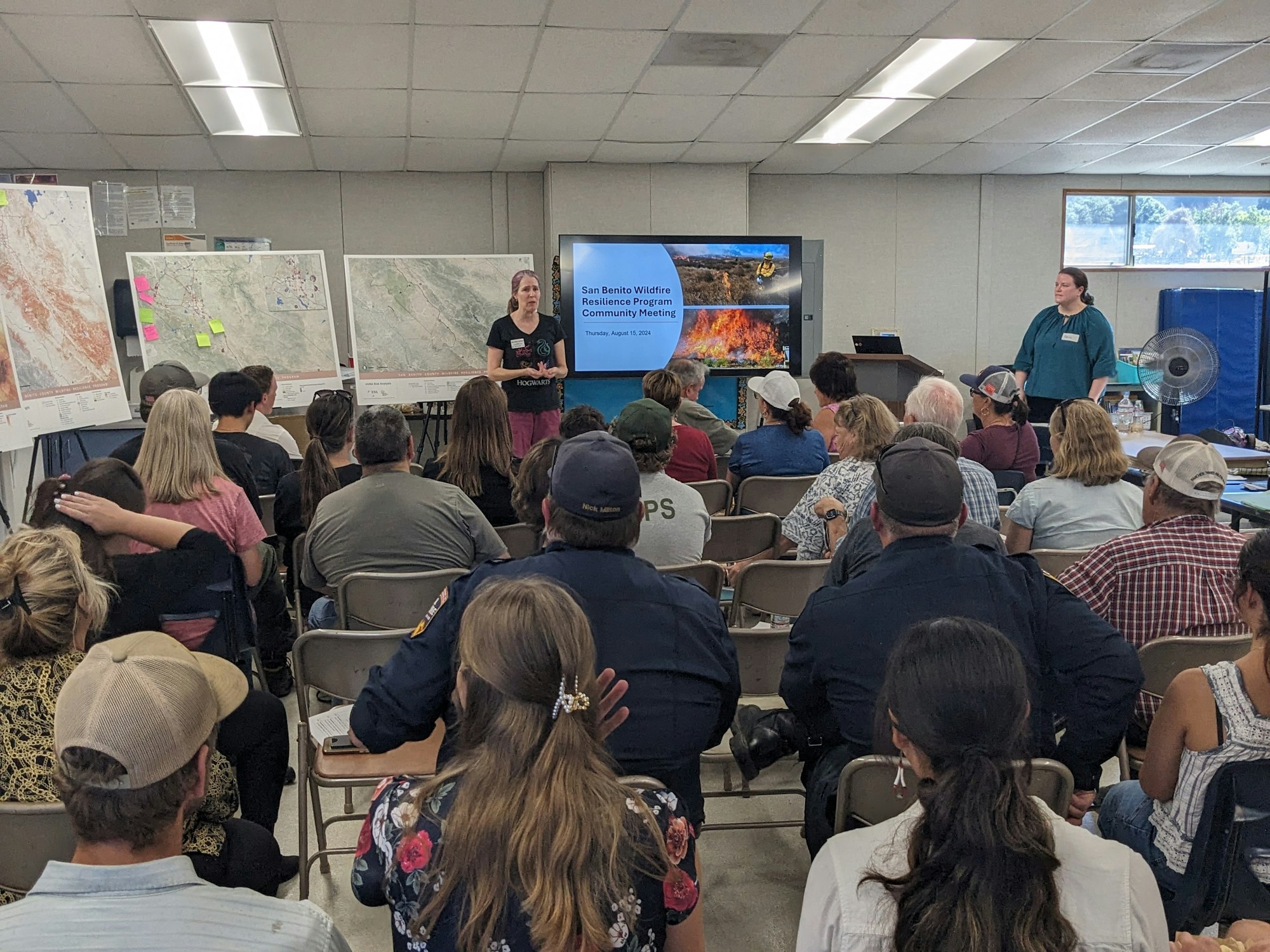 A community meeting with presenters and attendees, maps on display, and a slide about wildfire resilience.