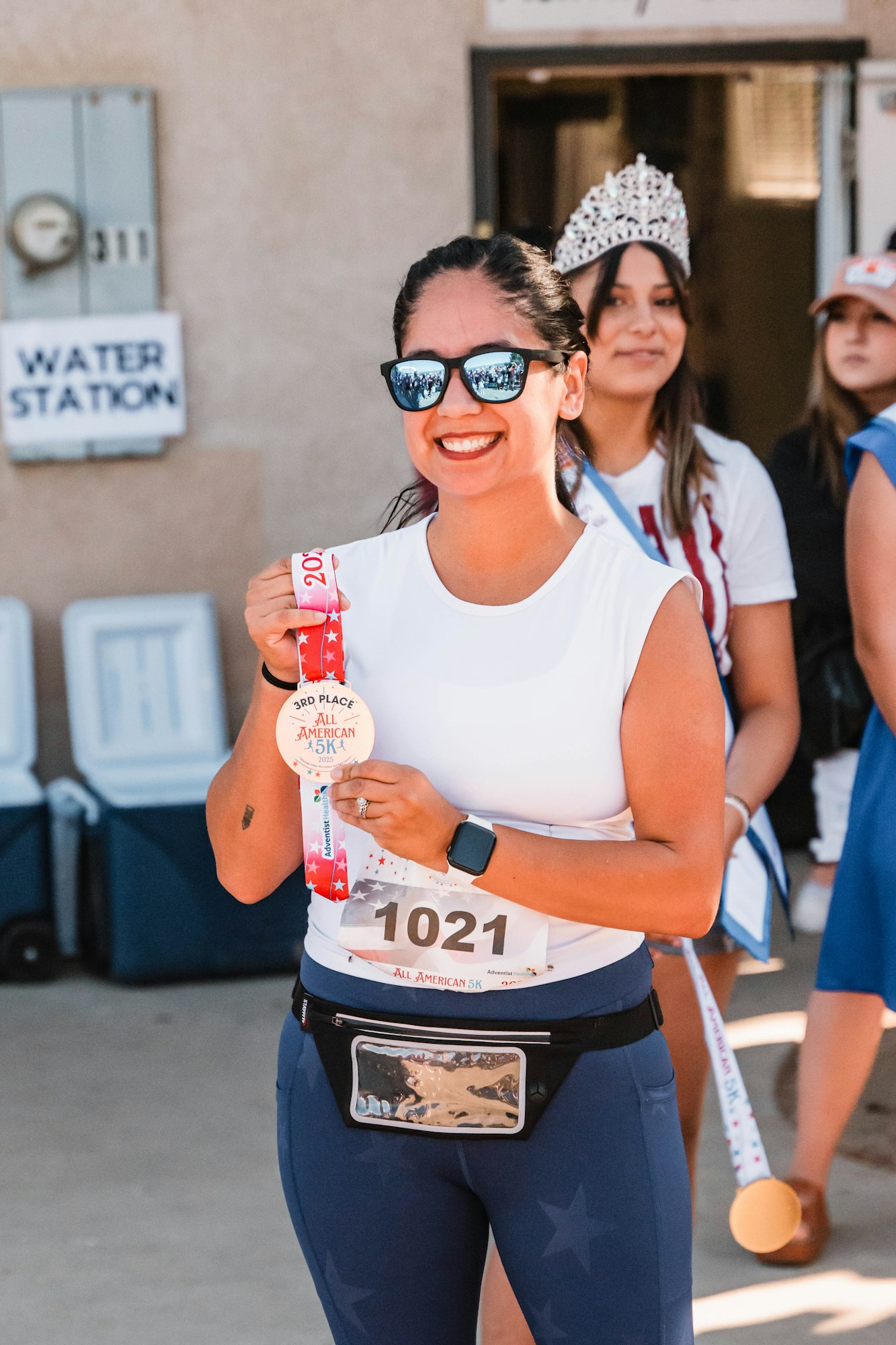 Woman holding a 3rd place medal for an "All American 5K" race, smiling, wearing sunglasses.