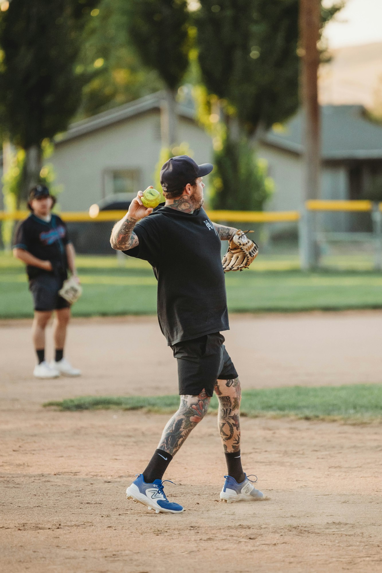 A tattooed player is throwing a softball on a field; another player is in the background.