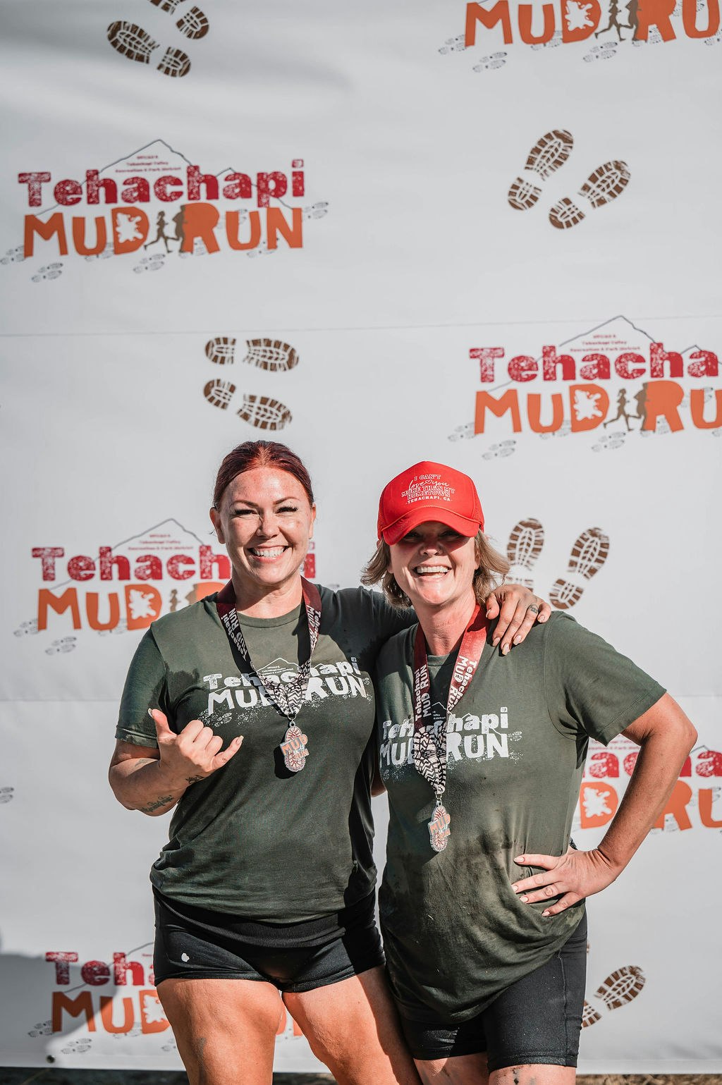 Two people smiling with medals in front of a "Tehachapi Mud Run" banner.