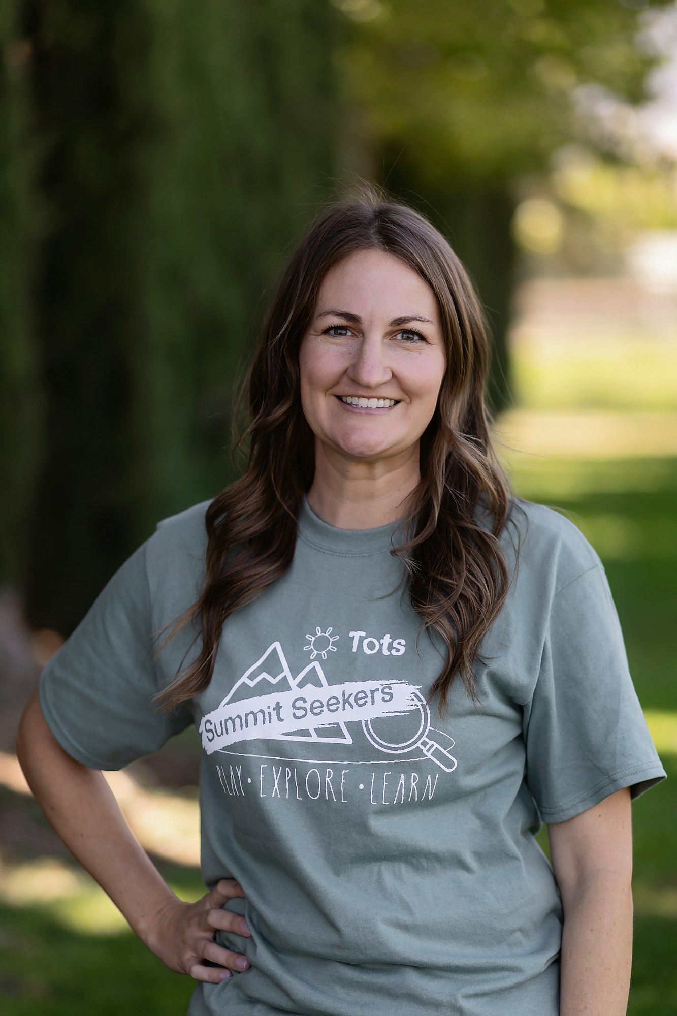A person in a gray "Summit Seekers" T-shirt is standing outside, smiling with trees and grass in the background.