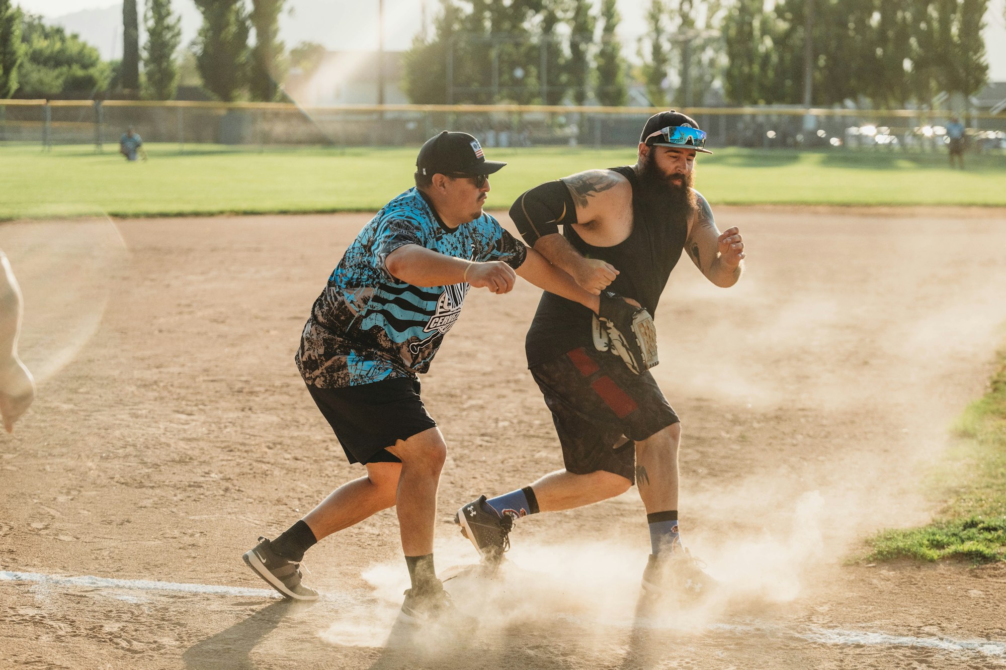 Two men playing softball, one running bases, the other trying to tag him out, dust flying, sunny field backdrop.
