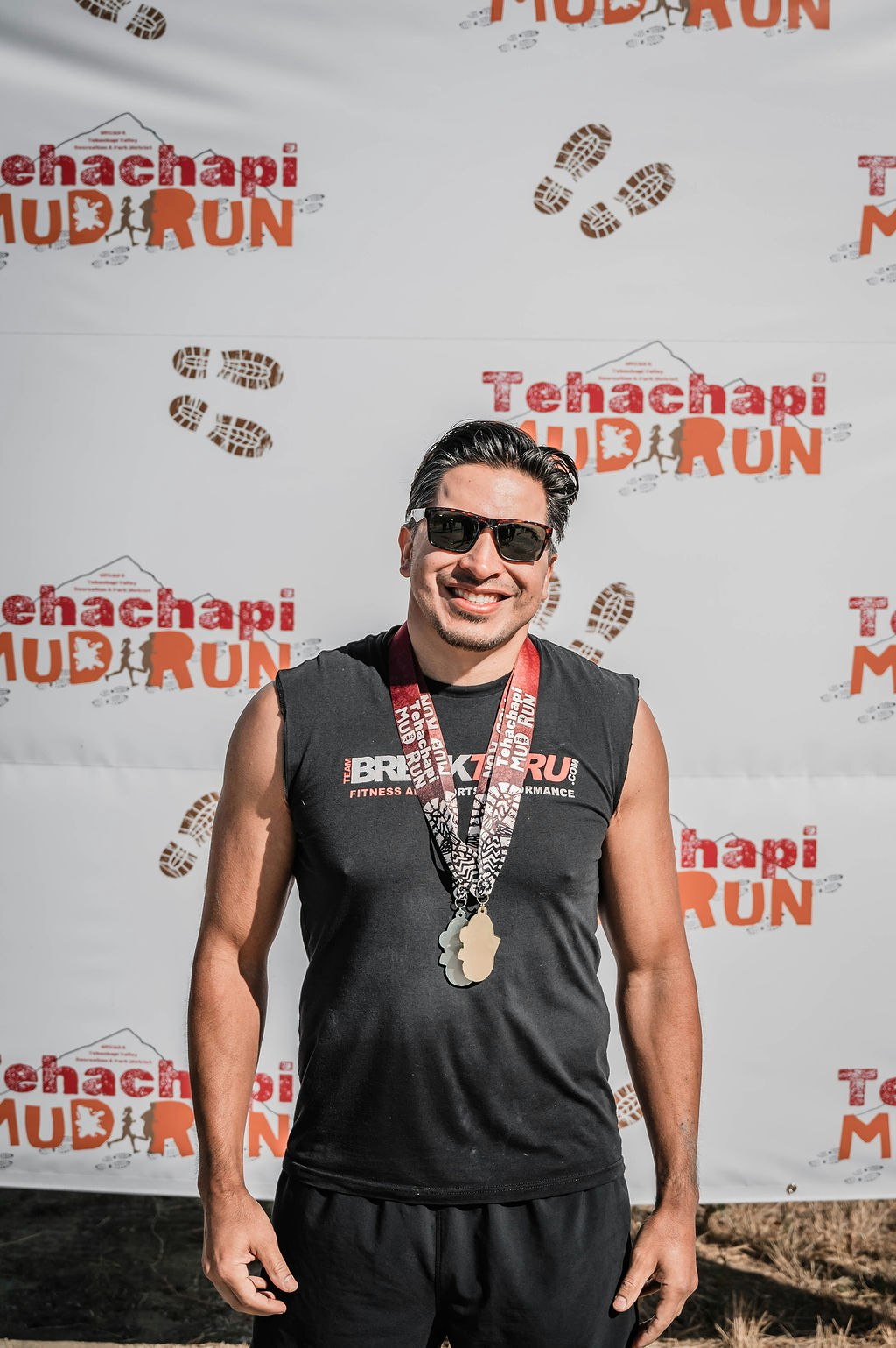 A person wearing a medal and sunglasses stands smiling in front of a "Tehachapi Mud Run" backdrop.