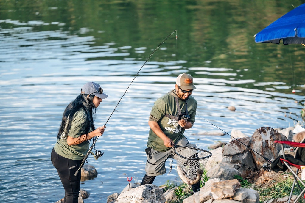 Two people enjoy fishing by a calm lake, one holds a fish net with a catch.