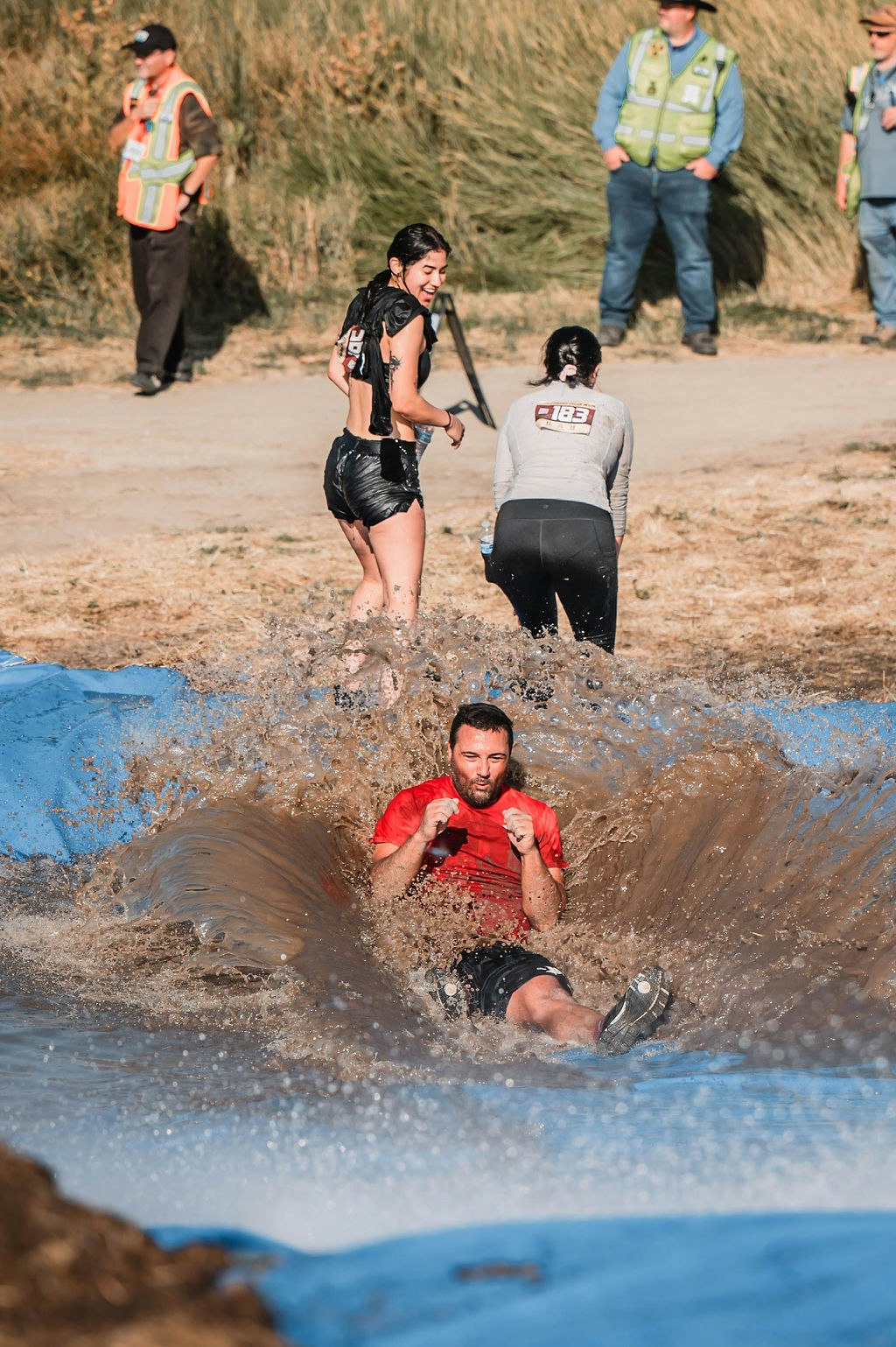 Man in red shirt slides into muddy water, woman laughs nearby, three people observe in the background.