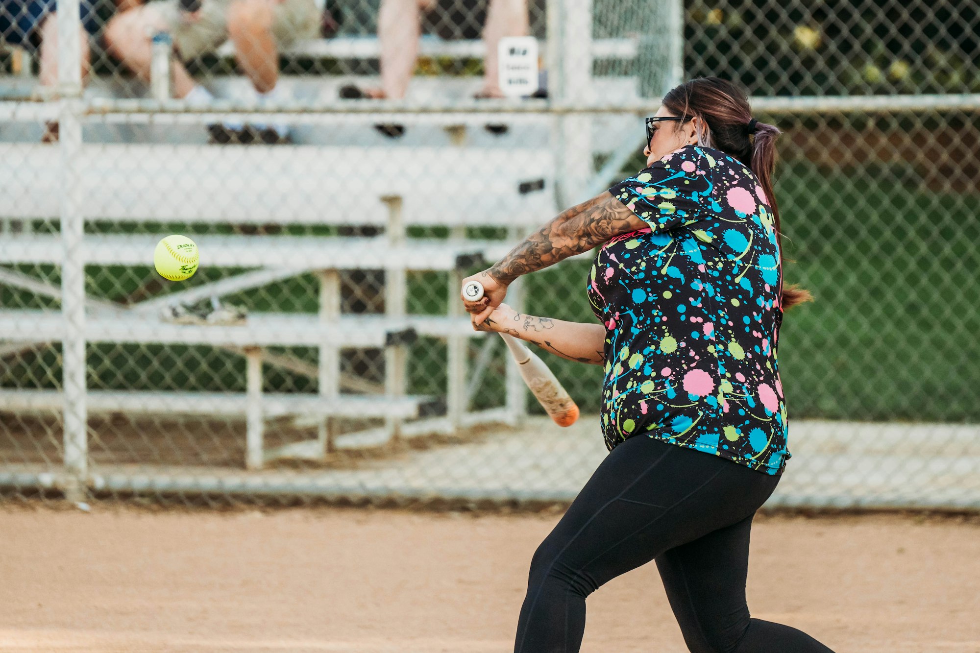A person swinging a bat at a softball with a chain-link fence and spectators in the background.