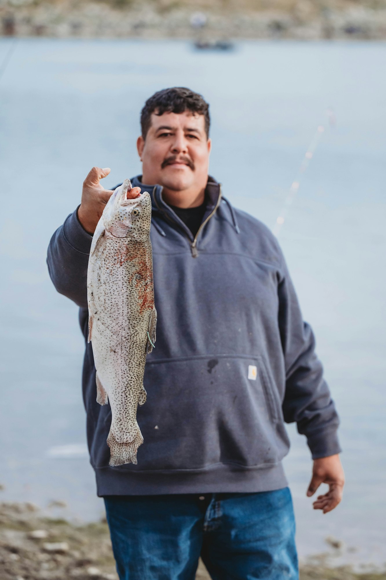A person holding a large fish near a body of water, wearing a dark pullover and jeans.