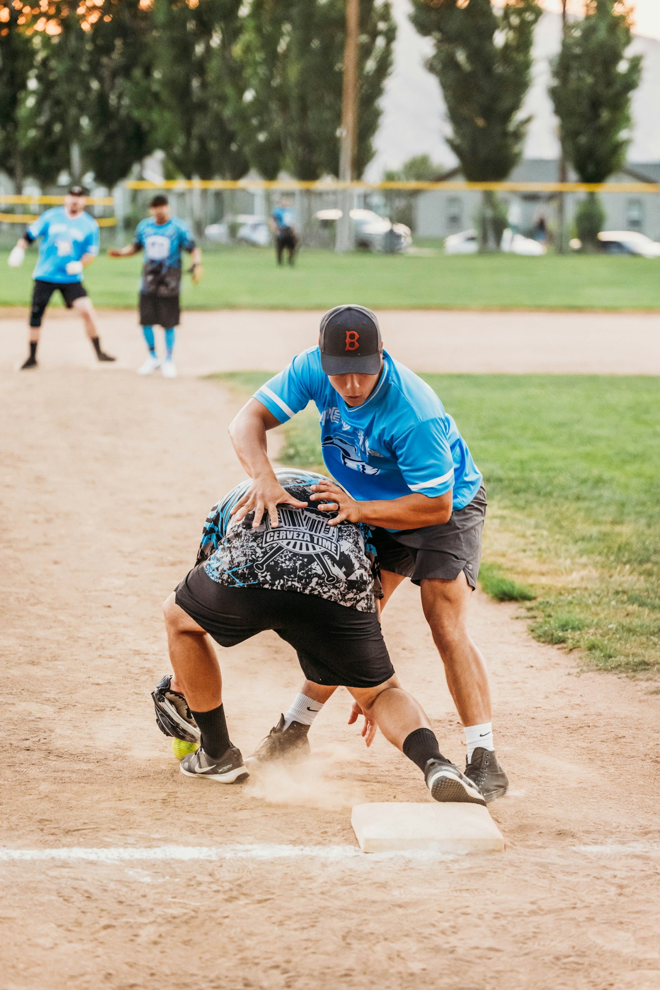 Adult softball game with a player sliding into base while another attempts to tag him out. Dust is kicked up, action-packed moment.