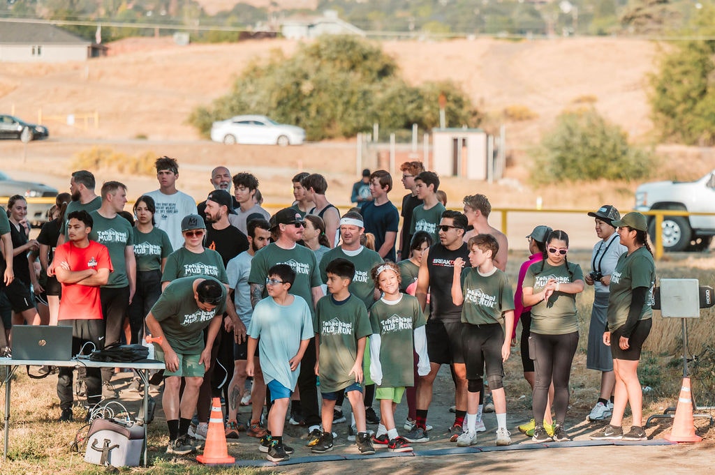 A group of people in sports attire, some wearing "Mud Run" shirts, gathered outdoors near a road with a laptop and cones.