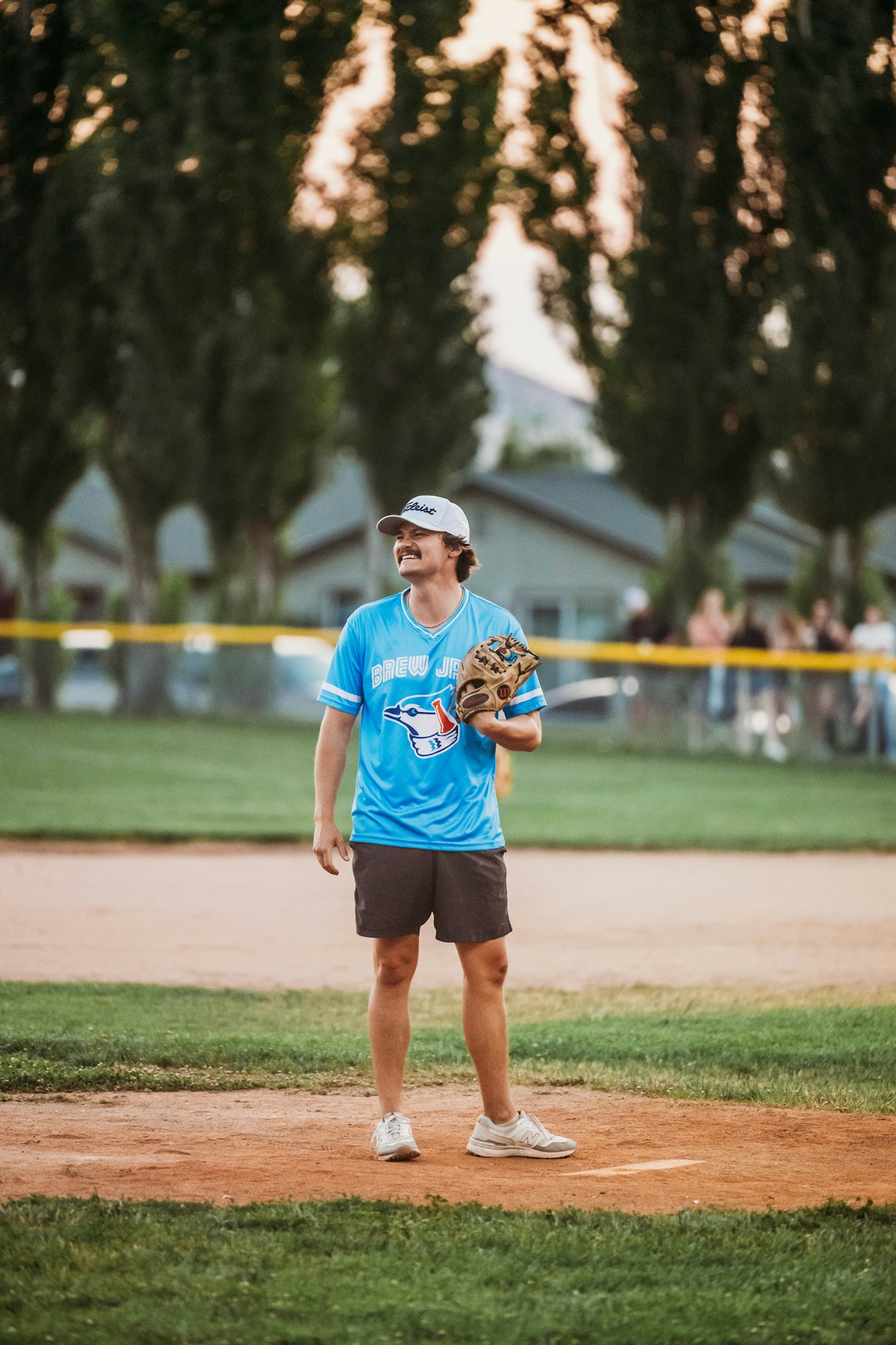 A person stands on a baseball field, wearing a glove, shorts, a blue jersey, and a cap.