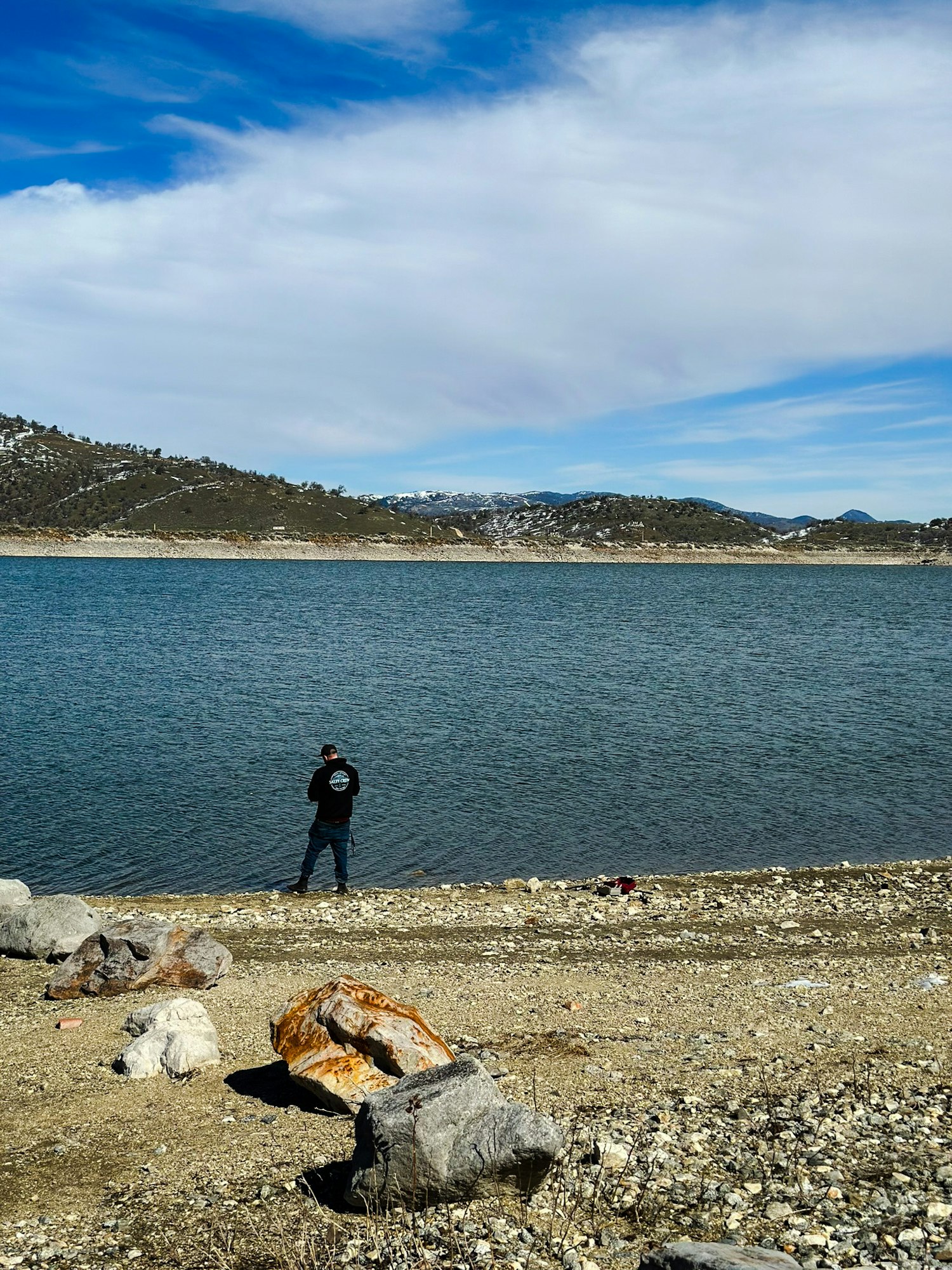 A person by a lake with mountains and partly snowy landscape under a blue sky with clouds. Rocks in the foreground.