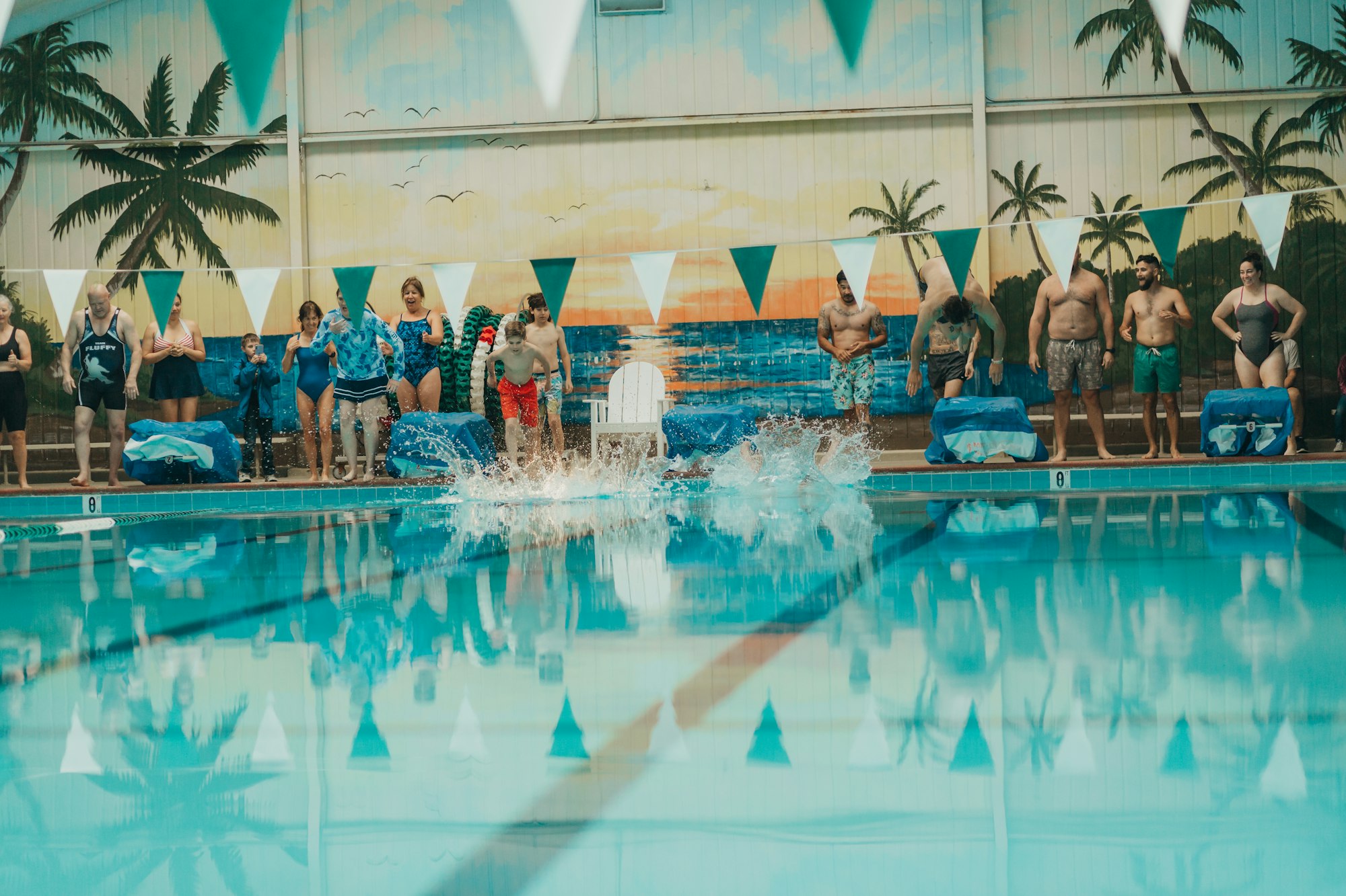 People at poolside, splash in water, indoor swimming pool with tropical mural, festive atmosphere.