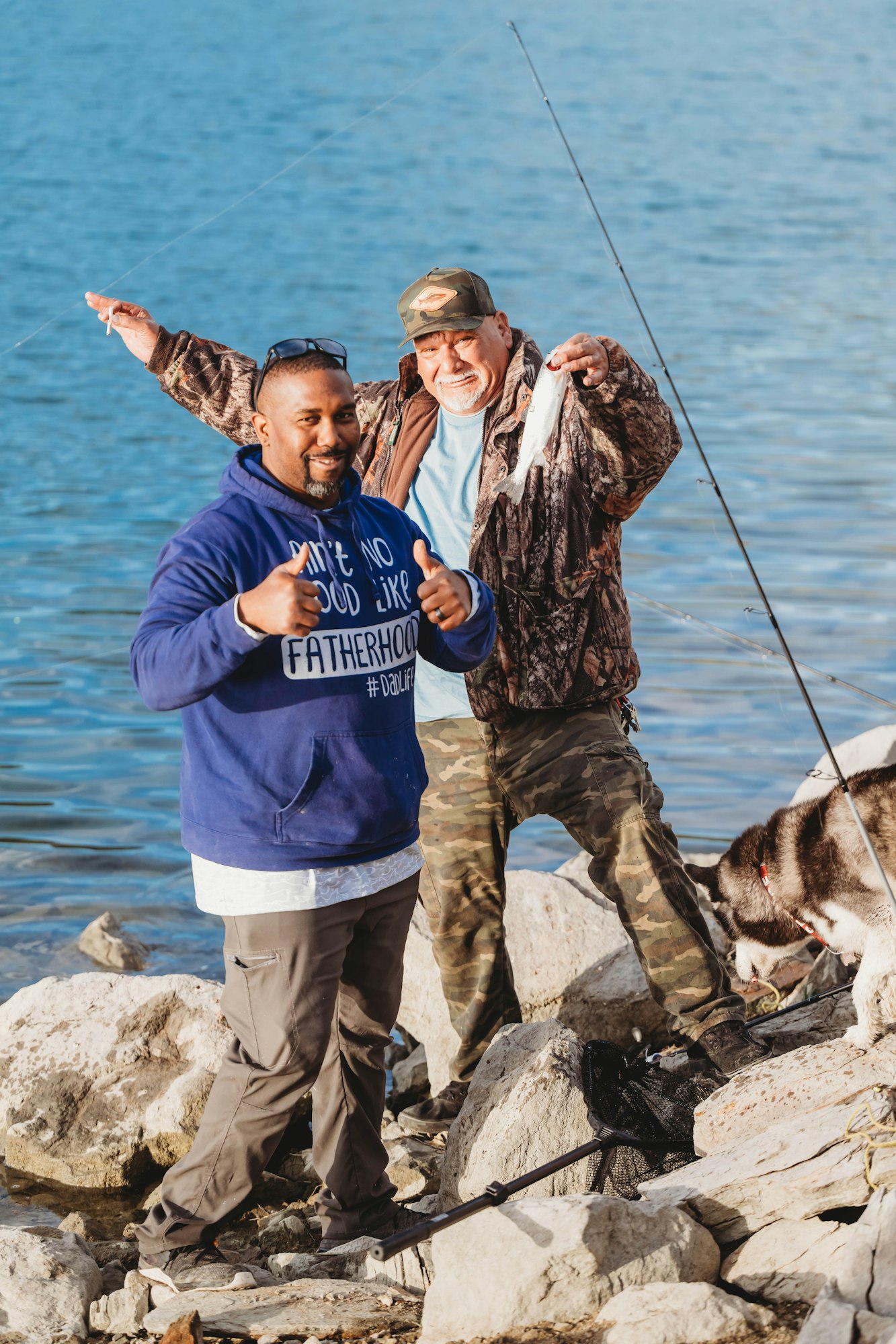 Two men fishing by a lake, one holding a fish, the other giving thumbs up. A dog is nearby on rocks.