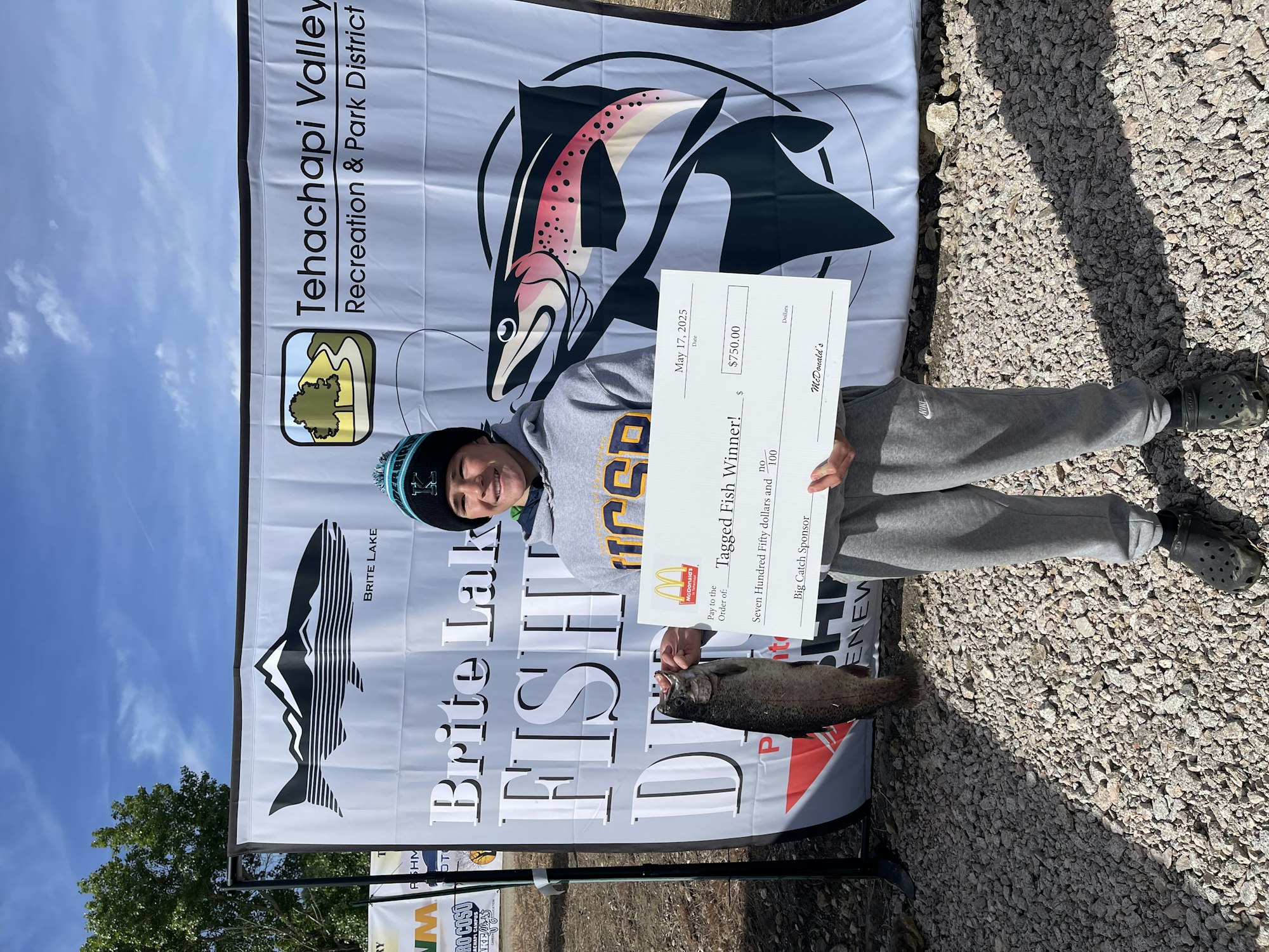 Person holding a large check and fish at a fishing event, with a "Brite Lake Fish" banner and logos in the background.
