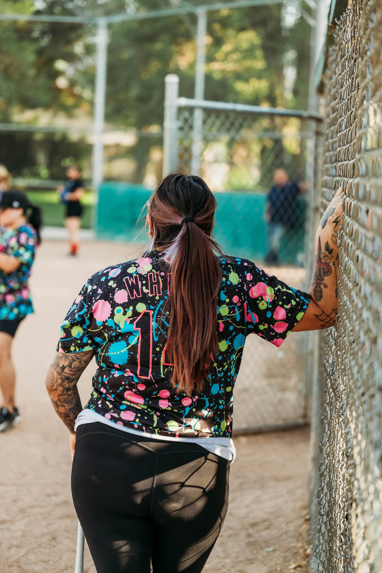 A person in a colorful sports jersey stands by a chain-link fence, watching others play in the background.